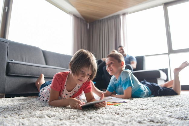Heureuse jeune famille jouant ensemble à la maison sur le sol à l'aide d'une tablette et d'un ensemble de dessins pour enfants