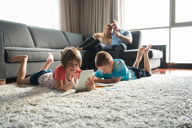 Heureuse jeune famille jouant ensemble à la maison sur le sol à l'aide d'une tablette et d'un ensemble de dessins pour enfants