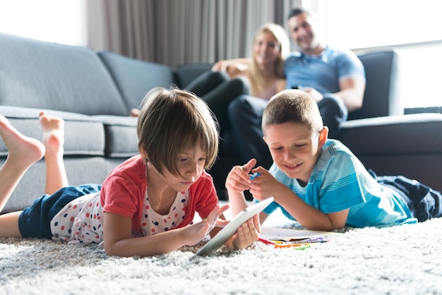 Heureuse jeune famille jouant ensemble à la maison sur le sol à l'aide d'une tablette et d'un ensemble de dessins pour enfants