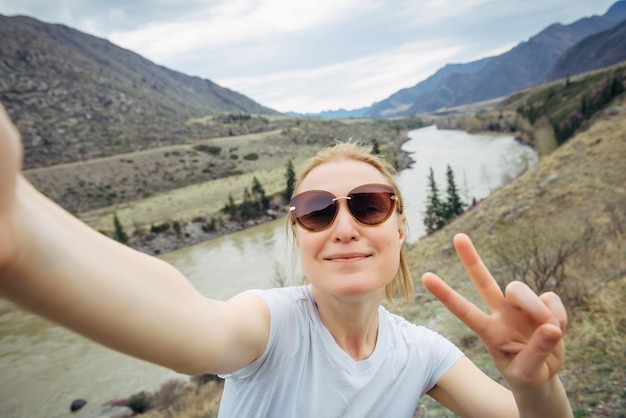 Heureuse jeune femme adulte en lunettes de soleil et T-shirt blanc prend un selfie, riant dans un appareil photo de smartphone contre le paysage de la rivière de montagne. Voyages, blogs, mode de vie actif.