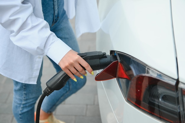 Heureuse jeune femme adulte souriante regardant loin en train de charger la batterie d'une petite station publique debout près d'une voiture électrique