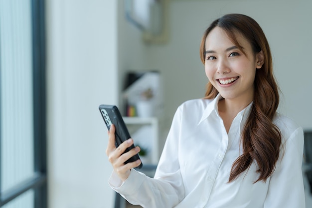 Heureuse jeune femme d'affaires asiatique portant un costume tenant un téléphone portable debout dans son bureau