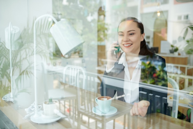 Heureuse jeune femme d'affaires buvant du café dans un café et parlant au téléphone avec un ami ou un collègue, vue de l'extérieur