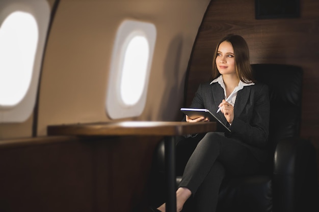 Heureuse jeune femme d'affaires caucasienne aux cheveux bruns tenant une tablette, assise sur un siège dans un jet privé, regardant la fenêtre de l'avion et souriant.