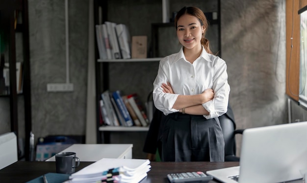 Heureuse jeune femme d'affaires confiante et détendue debout avec les bras croisés dans un bureau loft regardant la caméra avec un sourire chaleureux et amical