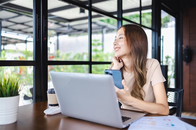 Heureuse jeune femme d'affaires gaie travaillant sur un smartphone et un ordinateur portable dans un café Technologie en ligne de travail et concept d'appareils modernes