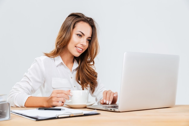 Heureuse jeune femme d'affaires séduisante buvant du café et utilisant un ordinateur portable sur fond blanc