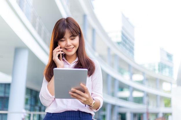 Heureuse jeune femme d'affaires séduisante dans sa chemise rose parlant sur son téléphone tout en utilisant sa tablette