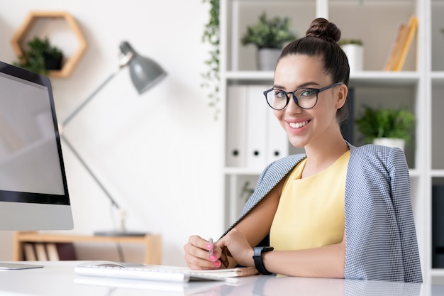 Heureuse jeune femme d'affaires en smart casual en vous regardant tout en étant assis par un bureau devant la caméra et la planification des travaux au bureau