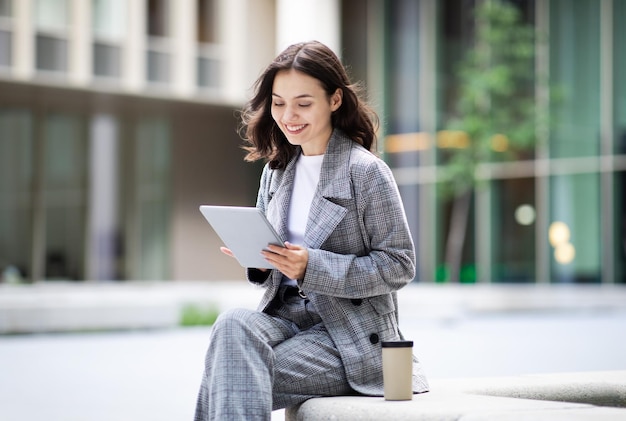 Heureuse jeune femme d'affaires tapant sur une tablette près d'un immeuble de bureaux