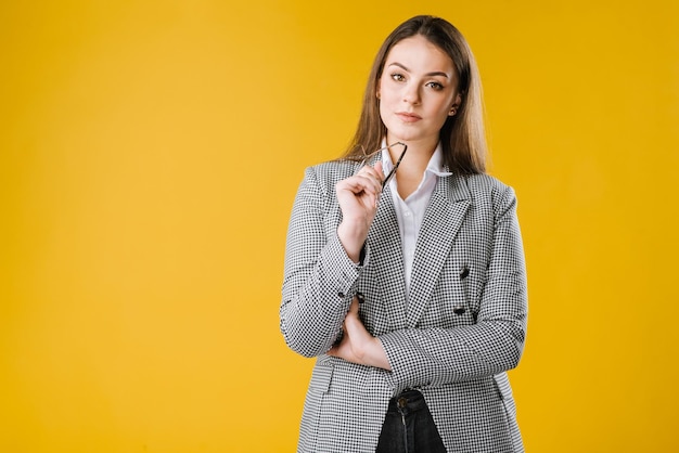 Heureuse jeune femme d'affaires en veste et chemise portant des lunettes sourire et posant devant la caméra sur fond jaune