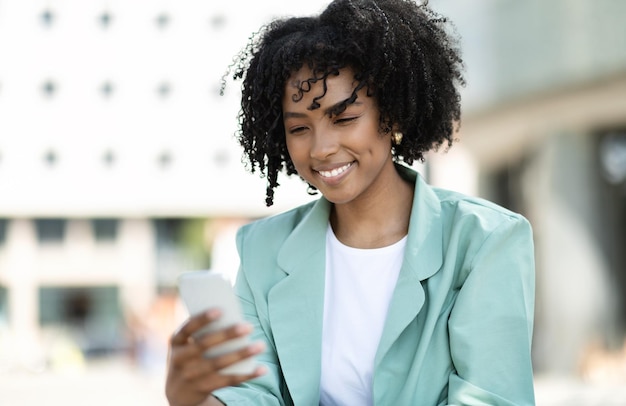 Heureuse jeune femme africaine gestionnaire utilisant le téléphone à côté de l'immeuble de bureaux