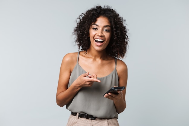 Heureuse jeune femme africaine habillée avec désinvolture debout isolé, tenant un téléphone mobile