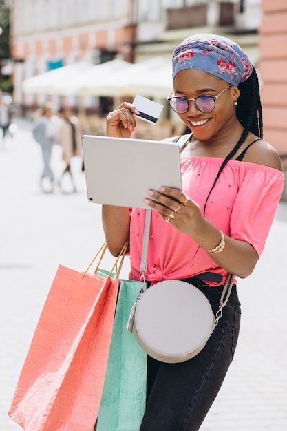 Heureuse jeune femme afro-américaine faire du shopping dans la tablette avec carte de crédit et tenant des sacs à provisions dans la rue