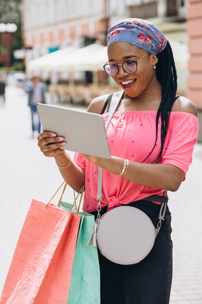 Heureuse jeune femme afro-américaine faire du shopping dans la tablette et tenant des sacs à provisions dans la rue