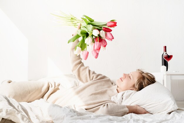 Heureuse jeune femme allongée dans le lit en pyjama tenant bouquet de fleurs de tulipes