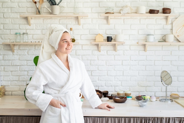 Heureuse jeune femme appliquant un gommage du visage sur son visage dans sa cuisine à domicile