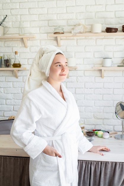 Heureuse jeune femme appliquant un gommage du visage sur son visage dans sa cuisine à domicile