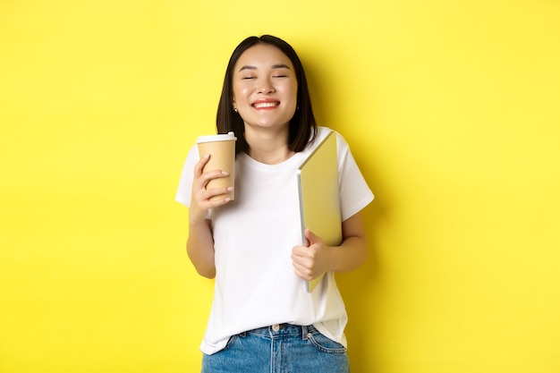 Heureuse jeune femme appréciant le café du café et souriant, tenant une tasse et un ordinateur portable, debout sur fond jaune