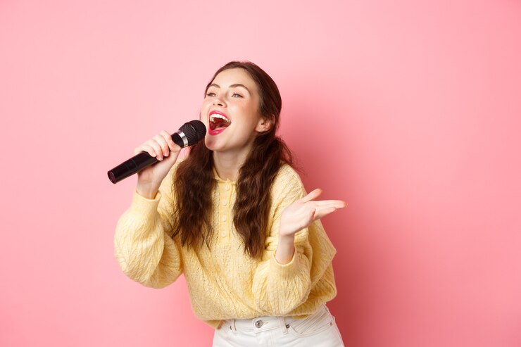 Photo heureuse jeune femme appréciant jouer au karaoké, chanter au micro, regardant de côté à l'écran avec des paroles, souriant joyeux, debout contre le mur rose.