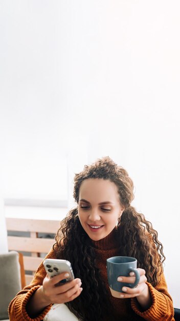 Heureuse jeune femme appréciant la technologie mobile tout en buvant du café à la maison souriante dame utilisant