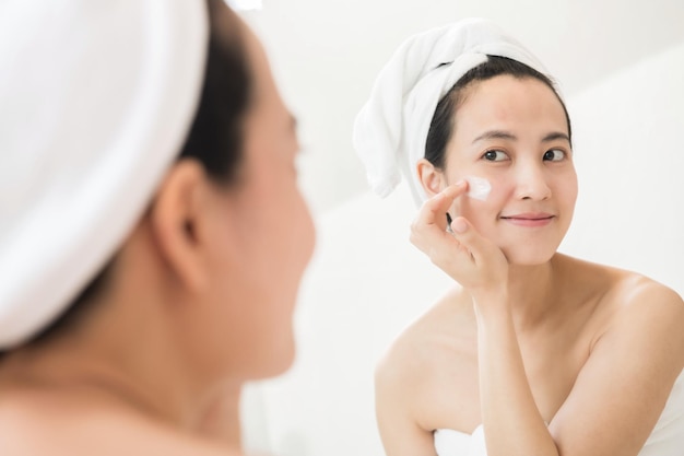 Heureuse jeune femme asiatique appliquant des lotions pour le visage tout en portant une serviette et en touchant son visage dans la salle de bain
