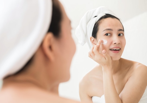 Heureuse jeune femme asiatique appliquant des lotions pour le visage tout en portant une serviette et en touchant son visage dans la salle de bain