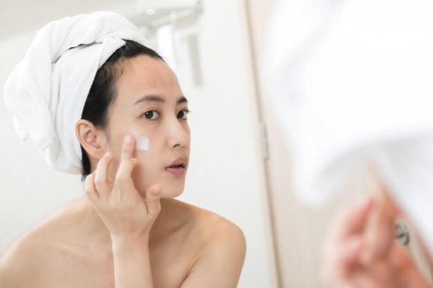 Heureuse jeune femme asiatique appliquant des lotions pour le visage tout en portant une serviette et en touchant son visage dans la salle de bain