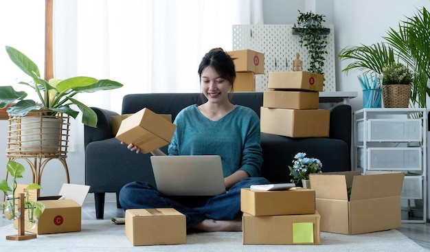 Heureuse jeune femme asiatique démarrage petite entreprise indépendante tenant une boîte à colis et un ordinateur portable et assis sur le sol Concept de livraison de boîte d'emballage de marketing en ligne