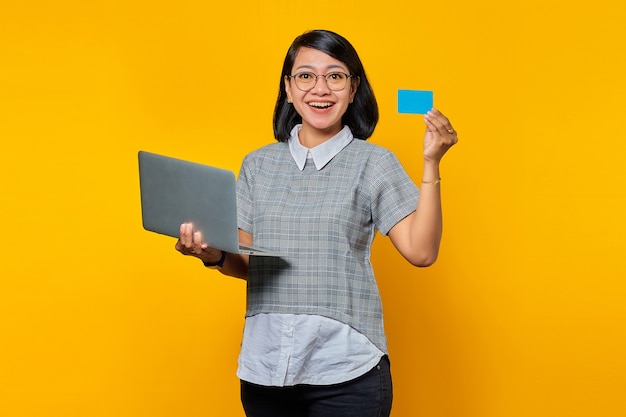 Heureuse jeune femme asiatique montrant une carte de crédit et un ordinateur portable à portée de main sur fond jaune