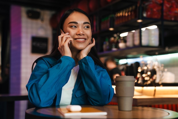 heureuse jeune femme asiatique parlant au téléphone portable et souriante assise dans un café chinois