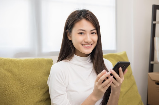 Heureuse jeune femme asiatique regardant l'écran d'un smartphone assis sur un canapé dans le salon, elle fait ses courses en ligne Elle est très heureuse