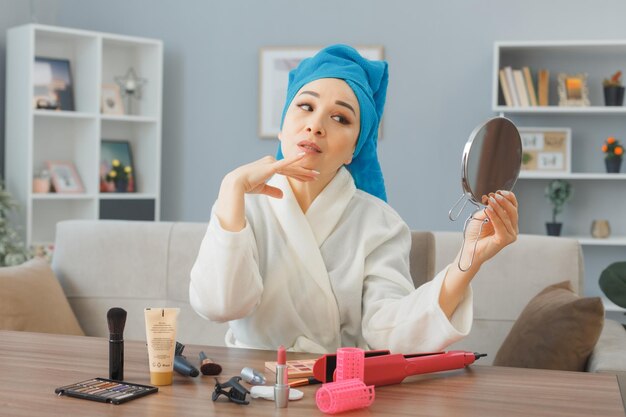 Heureuse jeune femme asiatique avec une serviette sur la tête assise à la coiffeuse à l'intérieur de la maison regardant un miroir faisant la routine de maquillage du matin