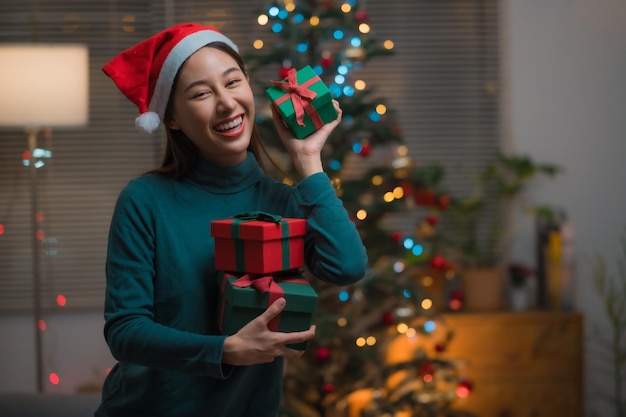 Heureuse jeune femme asiatique souriante en chapeau de père Noël tenant de nombreuses boîtes-cadeaux pour célébrer Noël et le nouvel an