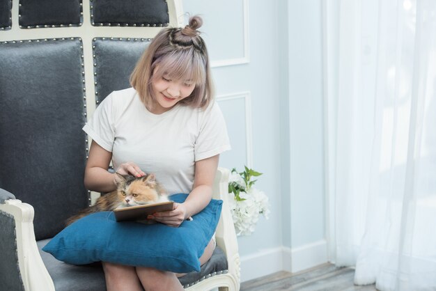 Heureuse jeune femme asiatique avec tablette dans ses mains et caresser chaton assis sur le canapé