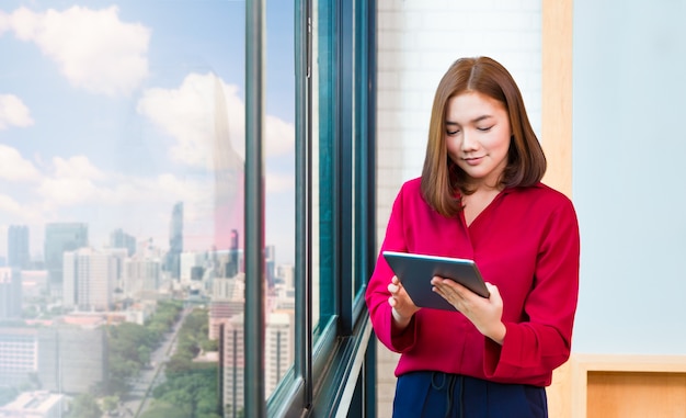 Heureuse jeune femme asiatique en utilisant sa tablette dans son bureau près de la fenêtre de grande taille