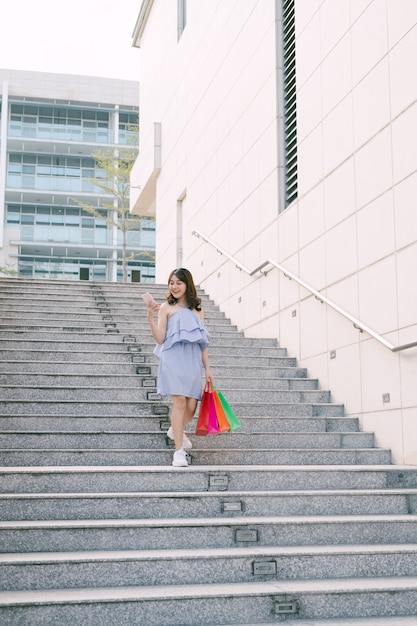 Heureuse jeune femme asiatique utilisant un téléphone et descendant des escaliers avec des emballages colorés.