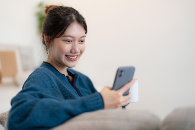 Heureuse jeune femme asiatique utilisant un téléphone portable tout en étant assise sur un canapé à la maison