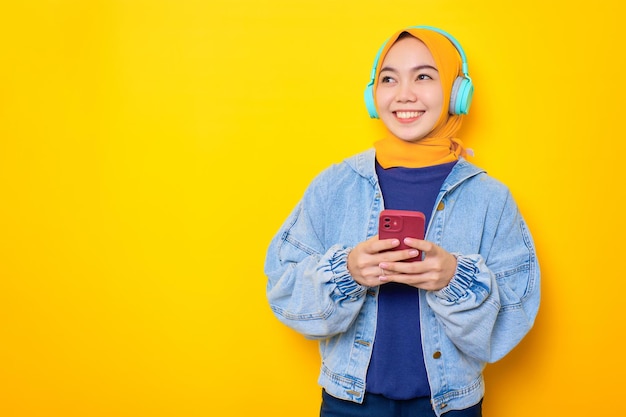 Heureuse jeune femme asiatique en veste de jeans et casque tenant un téléphone portable regardant de côté l'espace de copie isolé sur fond jaune