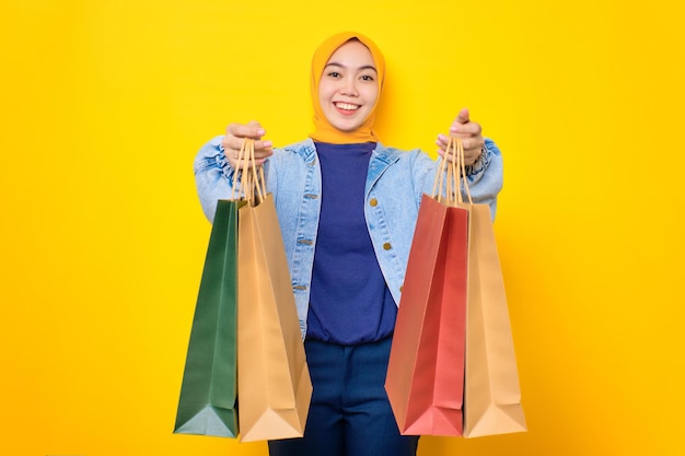 Heureuse jeune femme asiatique en veste de jeans tenant des sacs à provisions et souriant à la caméra isolée sur fond jaune