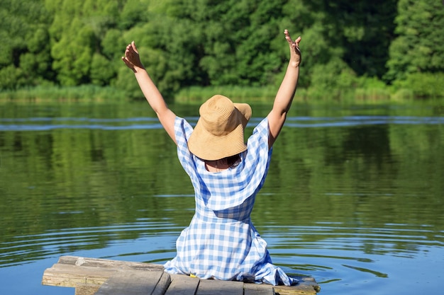 Heureuse jeune femme assise sur le pont près de la rivière Une robe bleue et un chapeau de paille Vue arrière