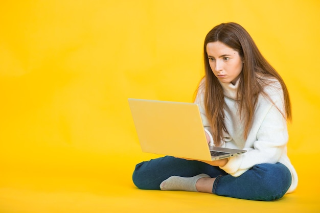 Heureuse jeune femme assise sur le sol avec les jambes croisées et utilisant un ordinateur portable sur jaune