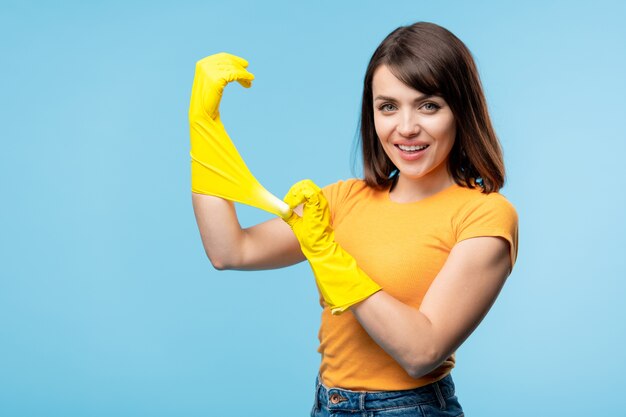 Heureuse jeune femme au foyer en t-shirt jaune mettant des gants en caoutchouc sur les mains avant de faire les tâches ménagères