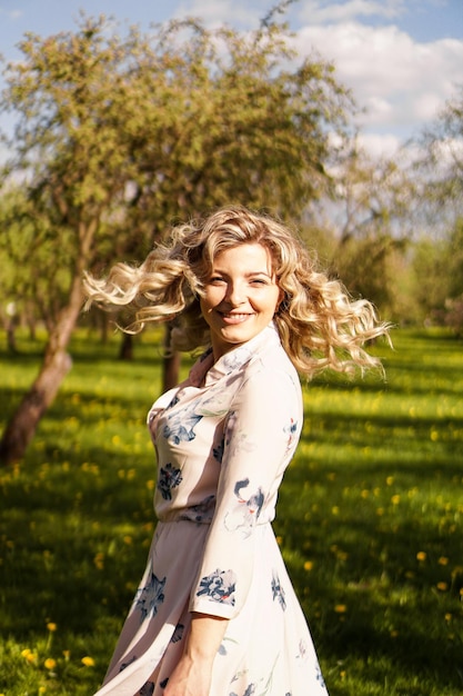 Heureuse jeune femme aux cheveux blonds, vêtue d'une robe, posant à l'extérieur dans un jardin avec des cerisiers au soleil, souriante. Boucles flottant au vent