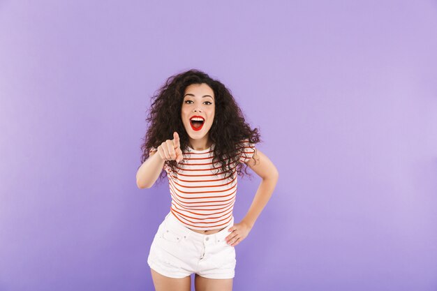 Heureuse jeune femme aux cheveux bouclés en été porter souriant et pointant le doigt sur vous