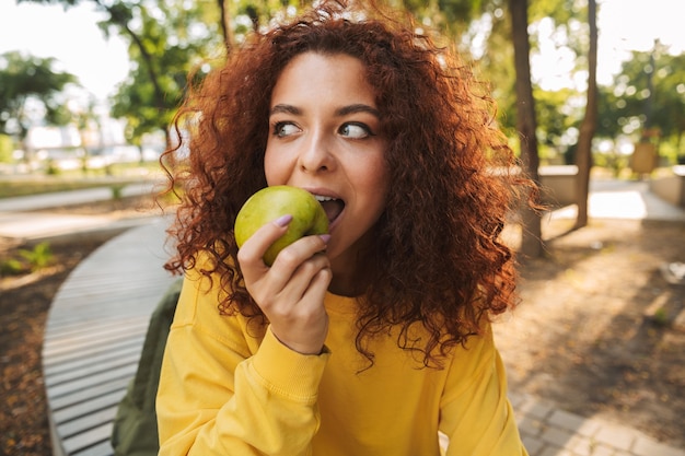 Heureuse jeune femme aux cheveux bouclés rouges assise sur un banc dans le parc, mordant la pomme verte