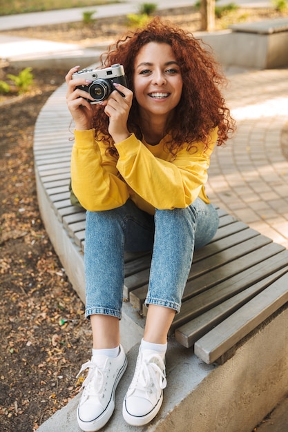 Heureuse jeune femme aux cheveux bouclés rouges assise sur un banc dans le parc, prenant des photos avec un appareil photo