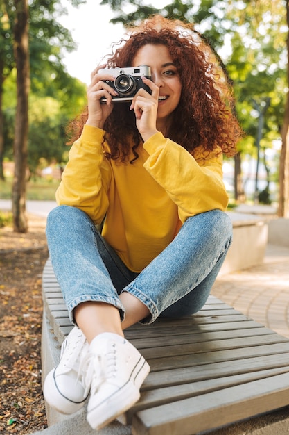 Heureuse jeune femme aux cheveux bouclés rouges assise sur un banc dans le parc, prenant des photos avec un appareil photo