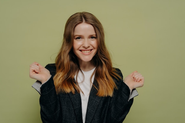 Heureuse jeune femme aux cheveux bruns ondulés tenant les mains en l'air démontrant l'excitation portant une veste formelle sombre et un haut blanc, posant sur fond de studio clair. Concept d'émotions humaines