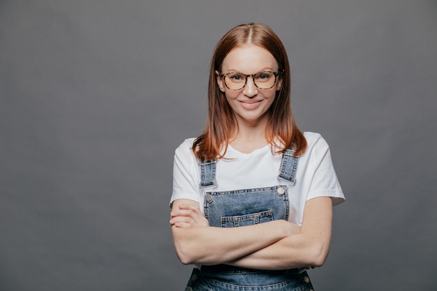 Heureuse Jeune Femme Aux Cheveux Noirs Garde Les Bras Croisés, Sourit Positivement, Porte Des Lunettes